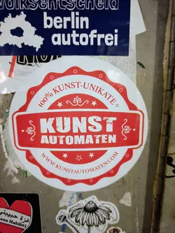 Street sticker A round sticker with a red central design element.  The text on the sticker reads "100% KUNST-UNIKATE" in a circular banner at the top, "KUNST AUTOMATEN" in the center, and "WWW.KUNSTAUTOMATEN.COM" at the bottom. The sticker promotes the website Kunstautomaten.com.
