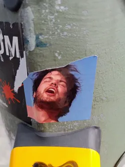A weathered sticker depicting a close-up of a man's face, his mouth open as if yelling or in pain. The background is a simple light blue sky.  The sticker is affixed to a textured, dirty surface.