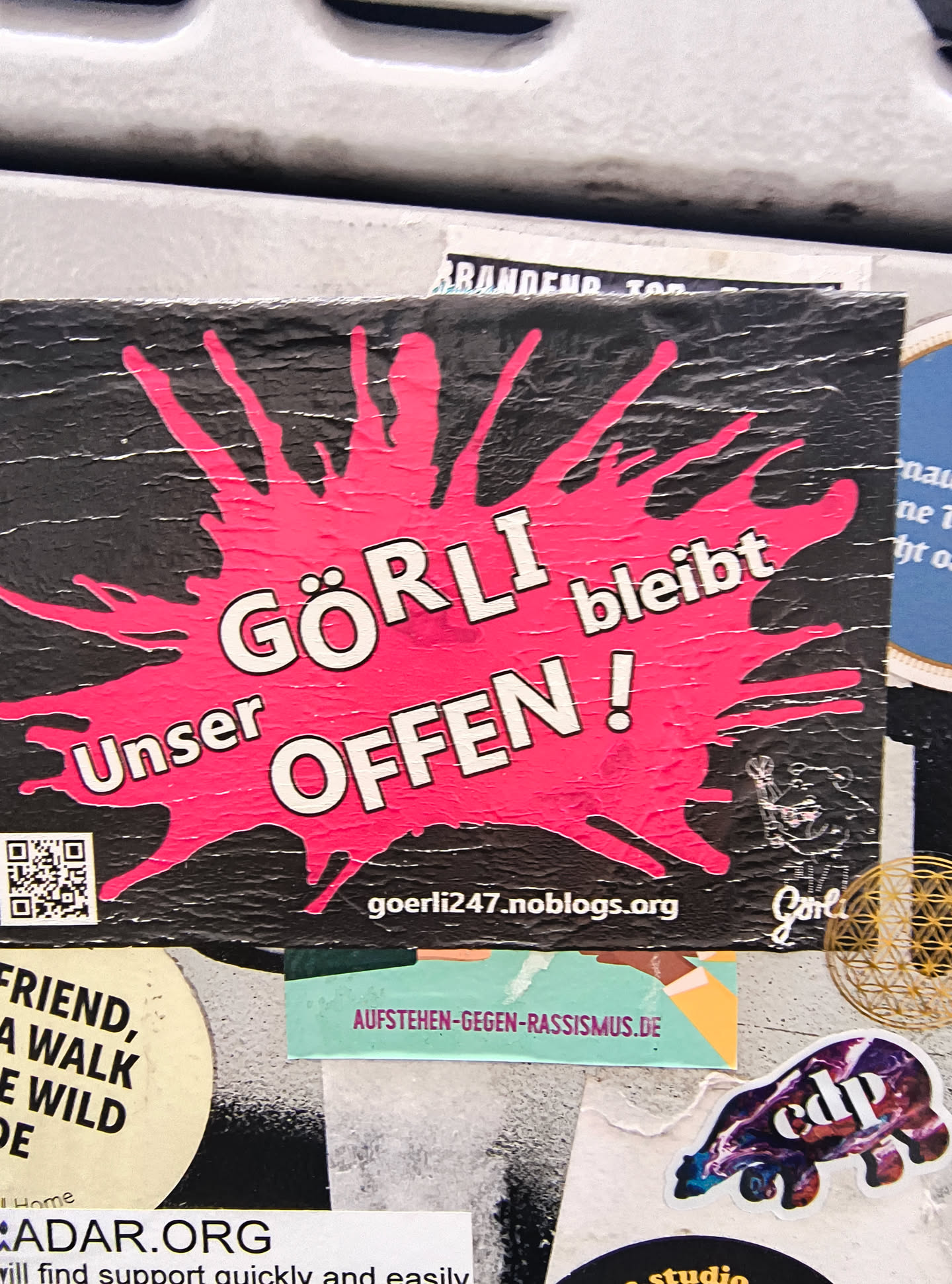 The sticker features a black background with a large, irregular splash of bright pink in the center. White, bold, sans-serif text is overlaid on the pink splash. The text reads 'GÖRLI bleibt' in the upper portion and 'Unser OFFEN !' in the lower portion, arranged in two lines. The letters have a subtle white outline. To the left of the text, there is a QR code. To the right of the text, there is a small, stylized graphic that appears to be a bear or a similar creature, and the word 'Görli' written in a script font. The sticker shows signs of wear and tear, with some creasing and texture on the surface.