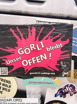 Street sticker The sticker features a black background with a large, irregular splash of bright pink in the center. White, bold, sans-serif text is overlaid on the pink splash. The text reads 'GÖRLI bleibt' in the upper portion and 'Unser OFFEN !' in the lower portion, arranged in two lines. The letters have a subtle white outline. To the left of the text, there is a QR code. To the right of the text, there is a small, stylized graphic that appears to be a bear or a similar creature, and the word 'Görli' written in a script font. The sticker shows signs of wear and tear, with some creasing and texture on the surface.