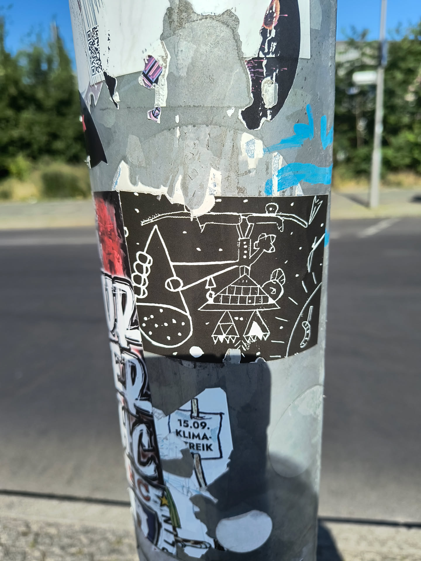This sticker on a pole shows a black background with a white doodle drawing. The drawing features abstract shapes, lines, and dots arranged in an odd and surreal composition. Some shapes resemble a house with a triangular roof and a hand holding a collection of circles. The style is whimsical and amateur, reminiscent of naive art. The surrounding pole has other stickers and markings, indicating a layered history of urban art.