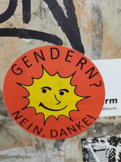 Street sticker A round sticker with an orange background and a yellow sun in the center. The sun has a slightly skeptical expression.  The text around the sun says "Gendern? Nein, Danke!" which translates from German to "Gendering? No, thank you!"