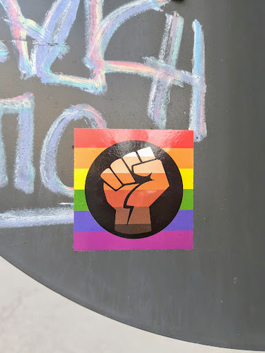 Street sticker The sticker features a rainbow pride flag with a raised fist in a black circle, showcasing support for LGBT rights and racial equality. The fist is gradient and shades of brown.