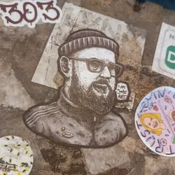 Street sticker This sticker features a stylized black and white illustration of a man's upper body. He has a beard, glasses, and is wearing a beanie and a zip-up jacket.  The style is reminiscent of printmaking or etching. Small text on the sticker says "I AM SHE ART".