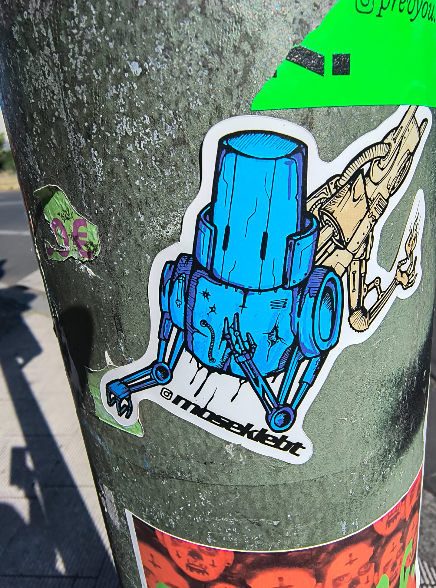 This is a sticker of a blue robot with a bucket-like head and two arms holding something. There is a golden firearm behind it. The text at the bottom of the sticker says "@moseklebt".