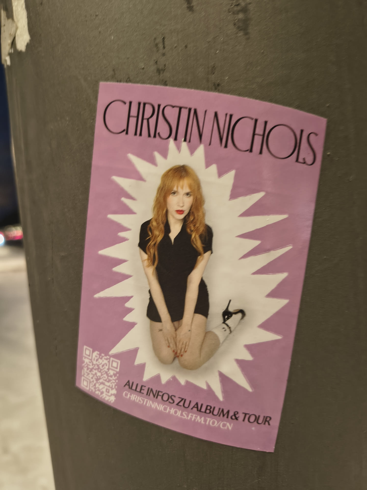 A rectangular sticker with a vibrant pink background, featuring a central image of a woman with long, wavy red hair and red lipstick, wearing a black collared shirt and black high heels with white socks. She is depicted in a kneeling or crouching pose, framed by a dynamic white starburst shape. Above her, the name 'CHRISTIN NICHOLS' is prominently displayed in black capital letters. Below the image, in black text, it reads 'ALLE INFOS ZU ALBUM & TOUR' followed by a short URL 'CHRISTINNICHOLS.FFM.TO/CN'. A scannable QR code is located to the left of this text.