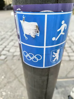 This sticker shows a design related to the Berlin Olympic Games and football. It features the Olympic rings, a football player pictogram, and a partially damaged BSC logo, likely representing a local sports club. The sticker is affixed to a pole outdoors, showing some wear and tear.