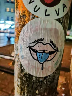 A round sticker featuring a cartoon drawing of a mouth with a tongue sticking out. The style is simple and slightly edgy, with a light pink mouth and a bright blue tongue. The sticker is affixed to a weathered pole, adding to its street art aesthetic. 
