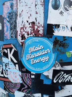 Street sticker A circular sticker with a bright, light blue background. The central text, 'Main Karakter Energy,' is written in a playful, bubbly script font with a white fill and a subtle light grey drop shadow, giving it a three-dimensional effect. Above this, curving along the top edge, are the words 'WE GIVE' in a smaller, simple white sans-serif font. Below the main text, 'EST. 2022' is placed similarly along the bottom curve. The sticker features two thin, concentric white lines forming a border just inside its perimeter. Its edges appear slightly worn and torn, revealing the texture of the surface it's adhered to, which is a distressed blue wall or pole covered with other layered posters and remnants.