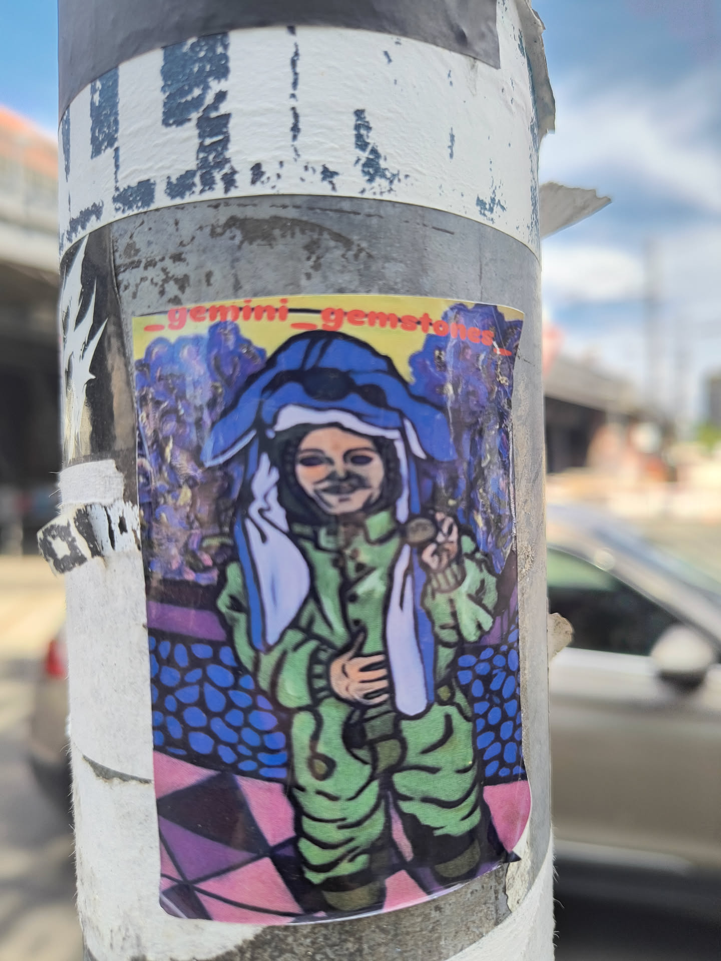 The sticker features a brightly colored, slightly abstract illustration of a character. The character is wearing a green jumpsuit and a large blue head covering that resembles an animal's ears and face. They have a subtle, enigmatic smile and are holding up a small, circular object in their right hand. The background is a psychedelic swirl of blues and purples, with a patterned floor below that transitions from blue ovals to pink and purple geometric shapes. The overall aesthetic is vibrant and whimsical, with a slightly surreal quality.