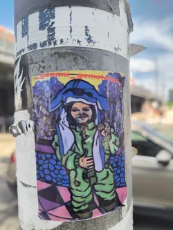 Street sticker The sticker features a brightly colored, slightly abstract illustration of a character. The character is wearing a green jumpsuit and a large blue head covering that resembles an animal's ears and face. They have a subtle, enigmatic smile and are holding up a small, circular object in their right hand. The background is a psychedelic swirl of blues and purples, with a patterned floor below that transitions from blue ovals to pink and purple geometric shapes. The overall aesthetic is vibrant and whimsical, with a slightly surreal quality.