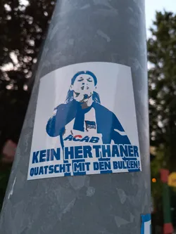 Street sticker A sticker on a metal pole shows a stylized image of a person with long hair, wearing a Hertha BSC soccer jersey with 'ACAB' written on it.  Beneath the image are the words, 'KEIN HERTHANER QUATSCHT MIT DEN BULLEN!' printed in blue.