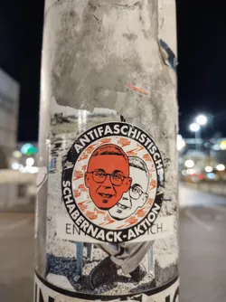 Street sticker A round sticker with an anti-fascist theme. It features two faces, one orange and one white, possibly representing different perspectives or individuals. The text on the sticker reads "Antifaschistisch Schabernack-Aktion..." which translates to "Anti-fascist prank action...". The sticker has a slightly distressed look, indicating it might be placed outdoors.