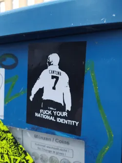 Street sticker A sticker featuring a stylized image of Eric Cantona, the famous Manchester United footballer, with his name and number 7 on the back of his shirt. The text 'Fuck Your National Identity' is printed below the image. The sticker is affixed to a blue metal surface.