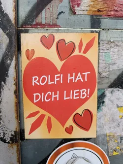 Street sticker A heart-shaped sticker with the text "Rolfi hat dich lieb!" (Rolfi loves you!) in German. The sticker has a light orange background and is decorated with several smaller hearts.