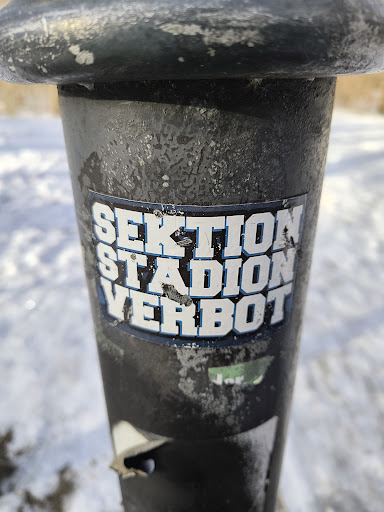 Street sticker The image shows a sticker affixed to a black metal pole. The sticker reads "SEKTION STADION VERBOT" in bold white letters with a blue outline. The pole is weathered with some chipped paint and surface wear. The background is blurred and shows a snowy or icy landscape.