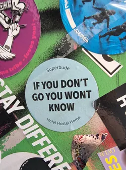 Street sticker A circular, light blue sticker with bold black text. The top of the sticker features the word 'Superbude' in a smaller font. The main text reads 'IF YOU DON'T GO YOU WONT KNOW' in large, uppercase letters. Below this, in a smaller, curved font, it says 'Hotel Hostel Home'. The sticker appears to be placed on a surface covered with other stickers and graffiti.