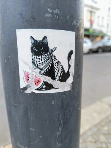 A sticker featuring a stylish black cat adorned with a keffiyeh scarf and enjoying a slice of watermelon. The sticker is slightly damaged, showing some peeling around the edges. It's affixed to a grey pole in an urban setting.