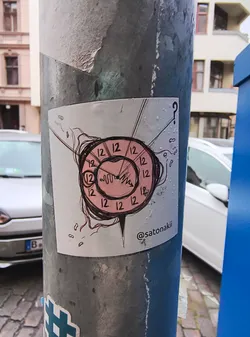Street sticker The sticker features a stylized clock face with a pink background and black outlines. The numbers '12' are repeated around the perimeter of the clock. The clock hands are drawn in a jagged, waveform-like style. Emanating from the clock are thin, dark lines that appear to be hair or tendrils, and a question mark is positioned above and to the right of the clock. The overall aesthetic is somewhat abstract and whimsical. Below the clock, a social media handle is visible.