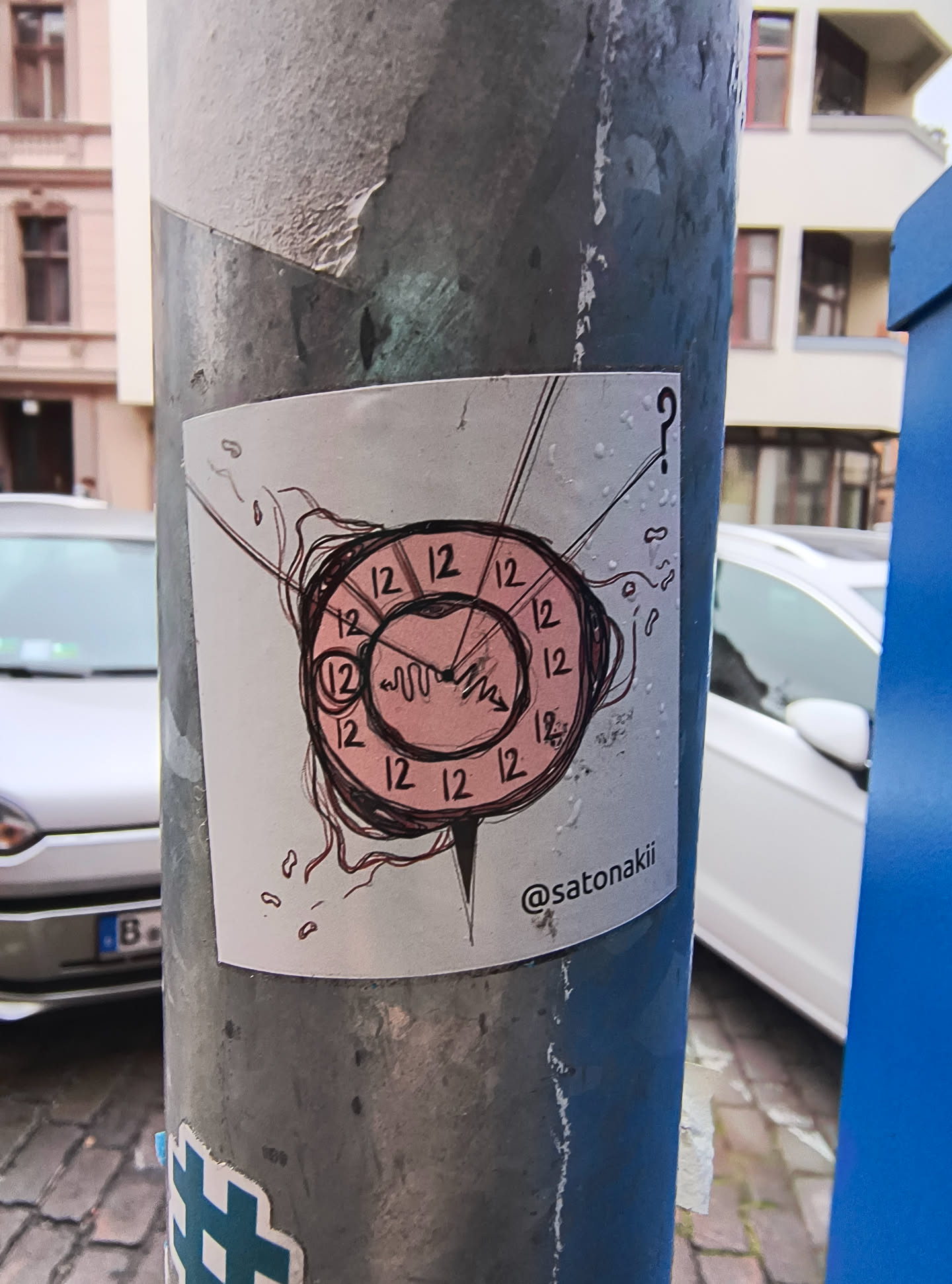 The sticker features a stylized clock face with a pink background and black outlines. The numbers '12' are repeated around the perimeter of the clock. The clock hands are drawn in a jagged, waveform-like style. Emanating from the clock are thin, dark lines that appear to be hair or tendrils, and a question mark is positioned above and to the right of the clock. The overall aesthetic is somewhat abstract and whimsical. Below the clock, a social media handle is visible.