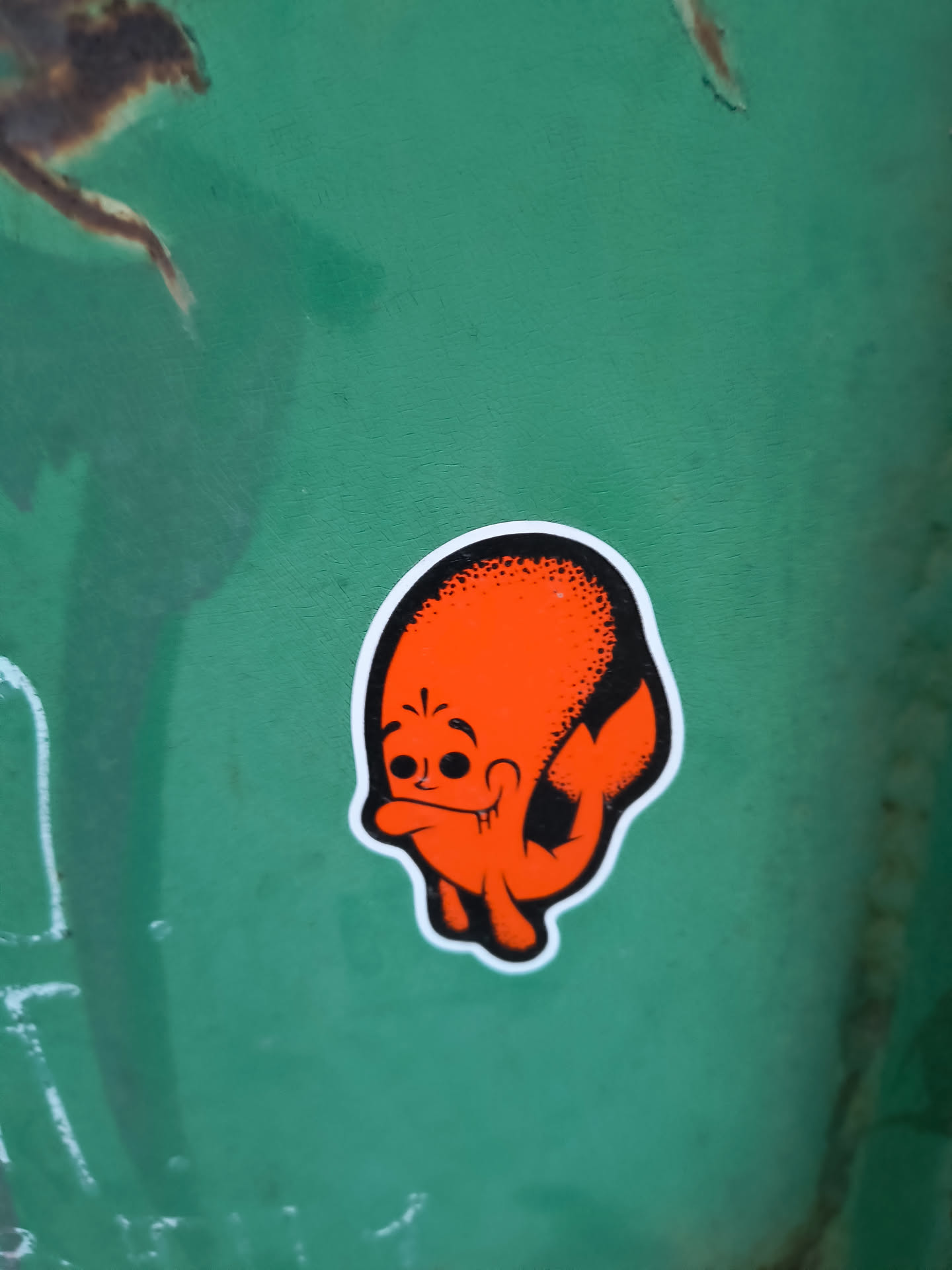 The sticker features a cartoonish character with an orange body and a black outline. The character has a large, round head with two black dots for eyes and a wide, smiling mouth with visible teeth. Its body appears somewhat amorphous, with two stubby arms and a tail-like appendage. The top of its head is textured with black stippling. The sticker has a white border.