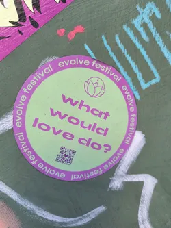 Street sticker This round sticker is light green with a purple border. The border has the words 'evolve festival' repeated around it.  In the center of the sticker are the words 'What would love do?' in purple, with a lotus flower symbol above the text and a QR code below. The sticker seems to be advertising Evolve Festival.