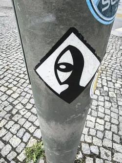 Street sticker The sticker is diamond-shaped, featuring a stark black and white design. The central image is a minimalist, abstract representation of a head or face, rendered in black silhouette against a white background. It has an elongated, somewhat hooded or alien-like profile. A prominent feature is a single, large, almond-shaped eye, which is white with a smaller black oval pupil, creating a striking contrast. The sticker has a thick black border outlining its diamond shape. The edges show some wear and slight peeling, indicating it's a physical sticker applied to the pole.