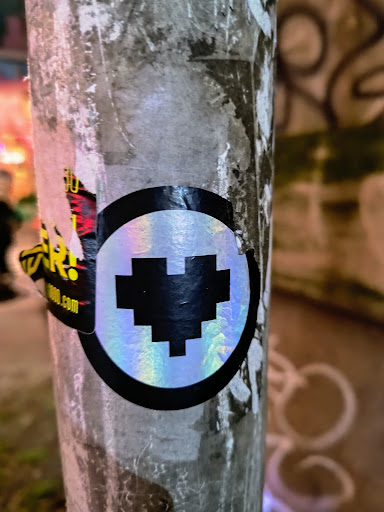 This sticker features a pixelated black heart enclosed in a holographic circle. It's pasted on a utility pole and is slightly damaged.