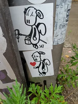 Two black and white stickers featuring an angel-like character. The top sticker shows the character holding a spray can and a spray nozzle. The bottom sticker shows the character holding a red heart.  Both stickers have a similar simplistic style.