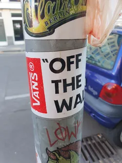 A classic Vans "Off The Wall" sticker is prominently displayed on a pole, amongst other stickers. The sticker is rectangular with red and white color scheme and features the Vans logo and the phrase "Off The Wall" in bold black lettering.