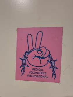 Street sticker The sticker is a rectangular design on a bright pink background. It features a central illustration in dark blue outline. The primary element is a hand making a peace sign (V-sign) with two fingers extended upwards. Wrapped around the wrist and extending downwards from both sides of the hand is a stylized depiction of barbed wire. Below the illustration, in dark blue capital letters, the text "MEDICAL VOLUNTEERS INTERNATIONAL" is displayed. The design is stark and graphic, using only two colors: vibrant pink for the background and dark blue for the illustration and text.