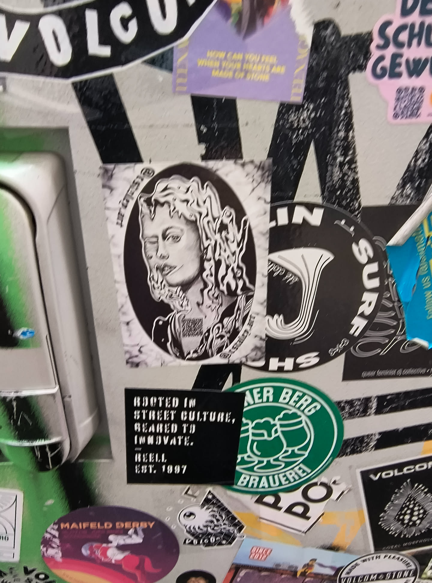 The sticker features a black and white oval illustration of a person's face. The person has wavy, almost liquid-like hair that frames their face. Their eyes are partially closed, and their lips are slightly parted. The style is detailed and somewhat surreal. There is a QR code visible on the chest area of the illustration. The sticker is surrounded by other stickers and appears to be on a textured surface.