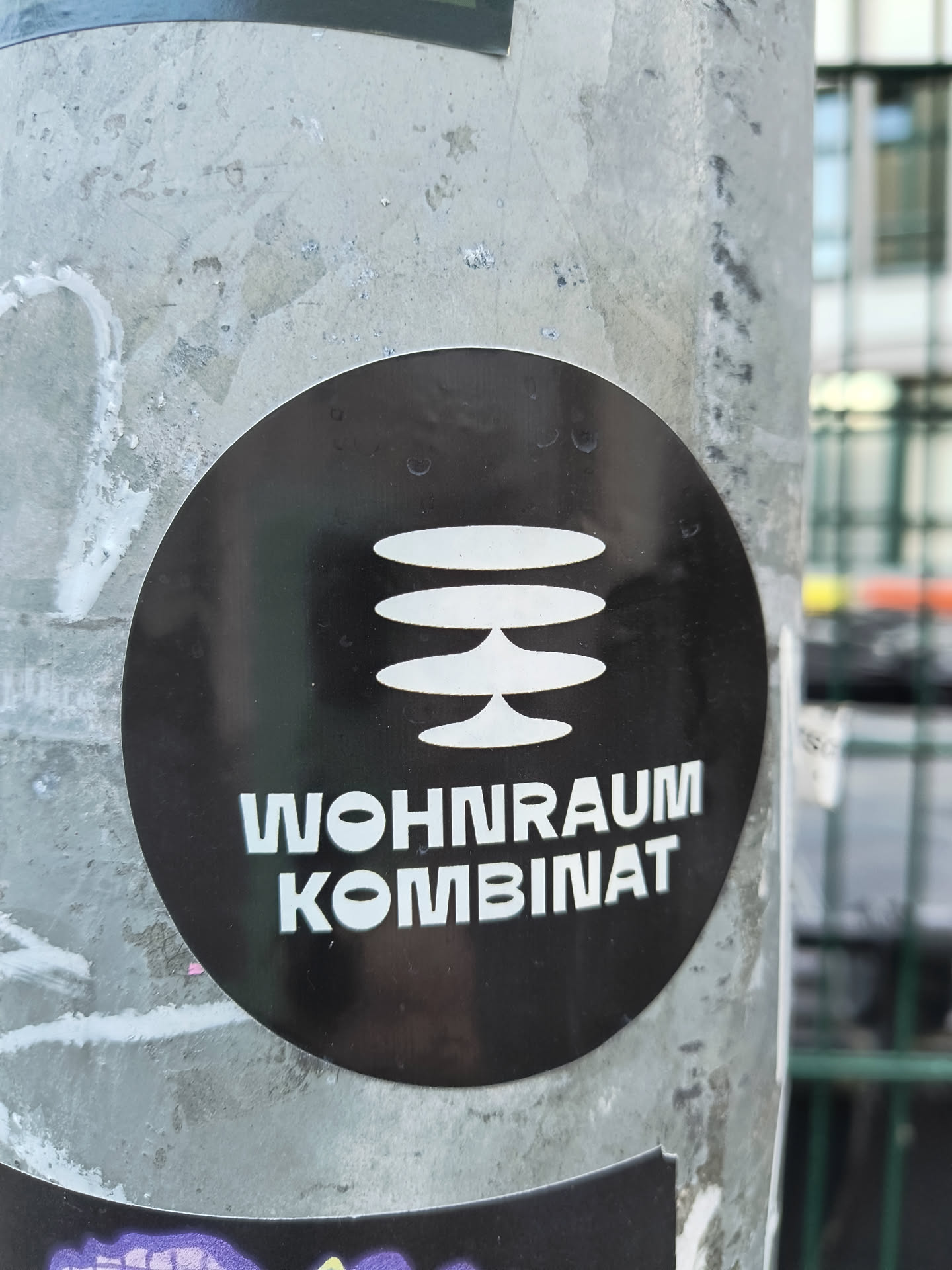 A round sticker with a black background and white text. The sticker has a symbol consisting of three horizontal lines stacked on top of each other, and connected to each other in the center, below the symbol are the words "WOHNRAUM KOMBINAT". The sticker is affixed to a gray metal pole.