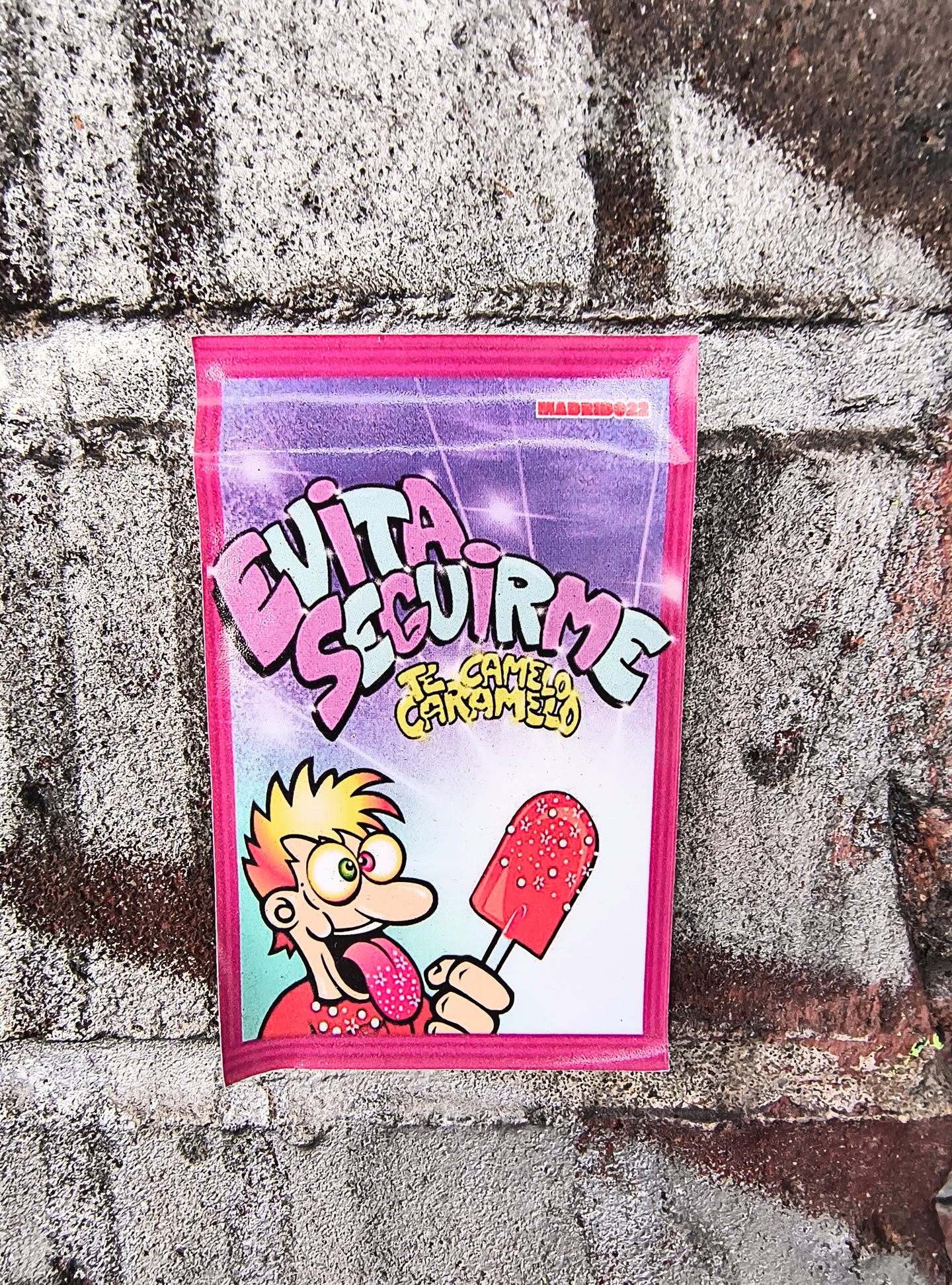 The sticker is predominantly pink with a purple background that has sparkling effects. In the center, large, bubbly text in light blue and pink reads 'EVITA SEGUIRME'. Below this, smaller yellow text says 'TU CARAMELO'. To the left, a cartoon character with spiky blonde hair, wide green eyes with pink irises, and an extended, patterned tongue is depicted. The character is wearing a red shirt with small white polka dots. The character is holding a red popsicle with white speckles and small pink flower-like designs on a stick. The sticker has a pink border.