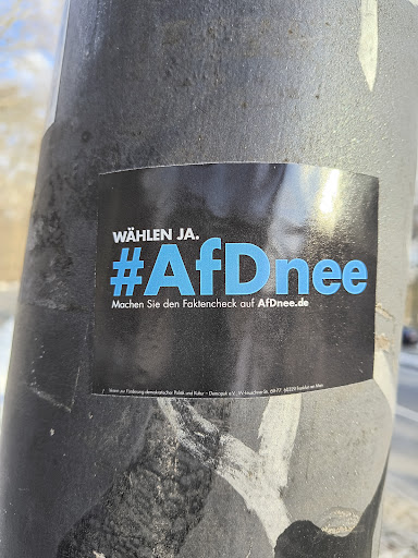 Street sticker A rectangular sticker is affixed to a gray pole. The sticker has a black background and blue text that reads 'WÄHLEN JA. #AfDnee Machen Sie den Faktencheck auf AfDnee.de'