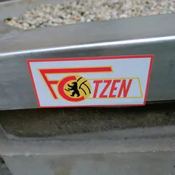 Street sticker A rectangular sticker featuring the logo of FC Kötzen. The logo has a red and yellow design with a bear emblem. The text 'FC' and 'KÖTZEN' is written in a bold font. The sticker is attached to a metal surface.