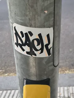 Street sticker A rectangular white sticker featuring a stylized black graffiti tag with faint peach-colored marker highlights, affixed to a galvanized metal pole.
