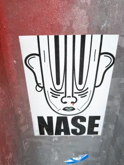 Street sticker The sticker is predominantly white with black line art. It depicts a face with large, prominent ears and a head that appears to be made of vertical black stripes. The facial features are simplified: small eyes, a nose with a single red droplet on the tip, and a small mouth. Below the face, the word 'NASE' is printed in large, bold, black sans-serif capital letters. The sticker has a slightly worn appearance, suggesting it has been applied outdoors.
