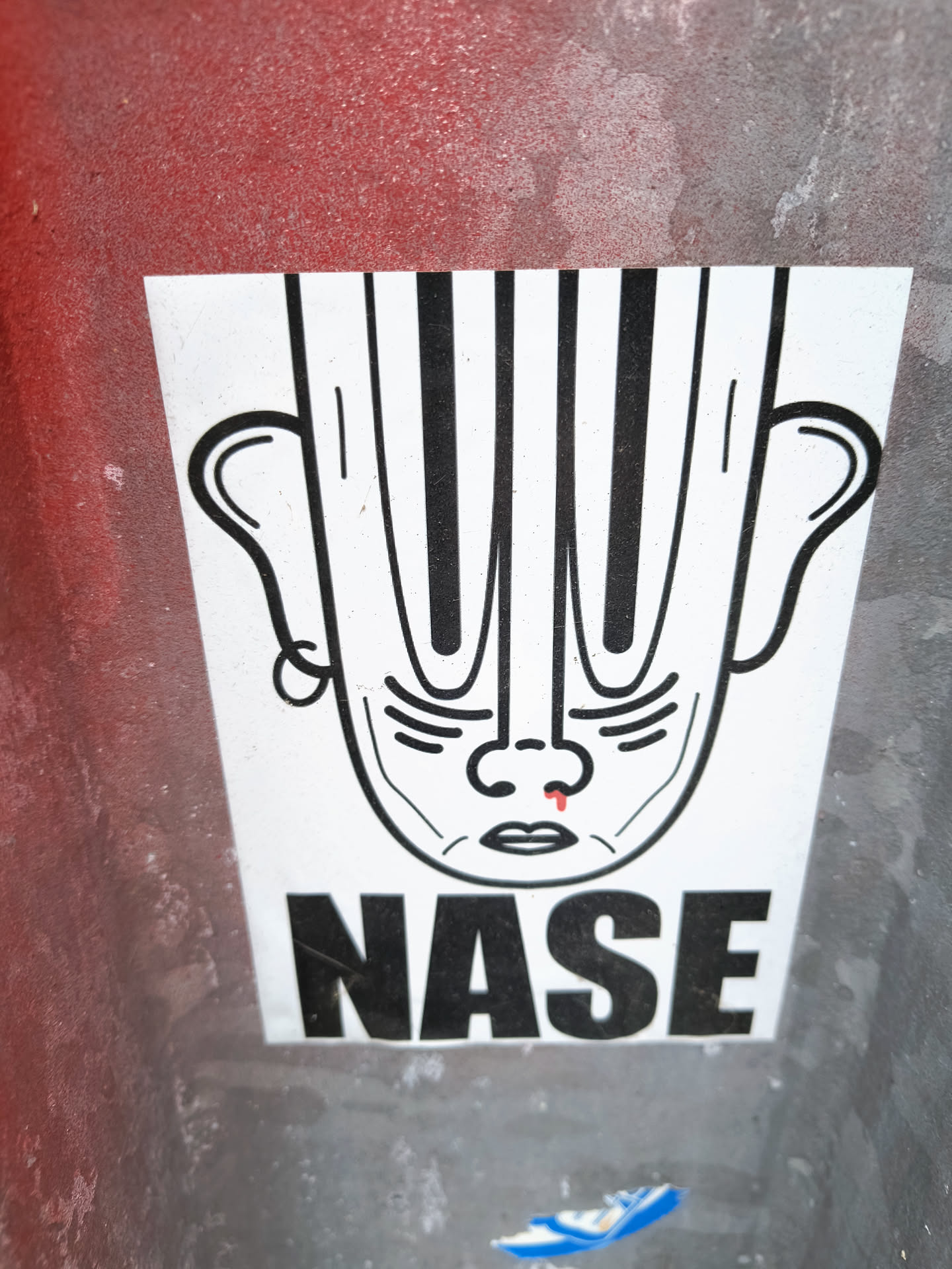 The sticker is predominantly white with black line art. It depicts a face with large, prominent ears and a head that appears to be made of vertical black stripes. The facial features are simplified: small eyes, a nose with a single red droplet on the tip, and a small mouth. Below the face, the word 'NASE' is printed in large, bold, black sans-serif capital letters. The sticker has a slightly worn appearance, suggesting it has been applied outdoors.