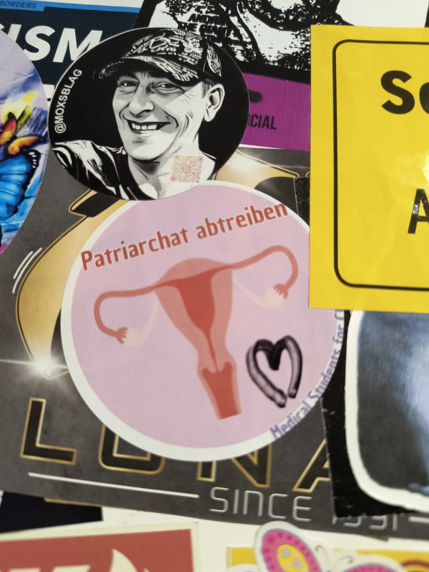 The central sticker is circular with a light pink background and a white border. It features a stylized illustration of a uterus in a gradient of coral pink to a lighter pink. To the right of the uterus, there is a dark grey heart shape with a textured, almost smudged appearance. Arching over the top of the sticker in reddish-brown text are the words 'Patriarchat abtreiben'. Along the bottom edge of the sticker, also in dark grey text, are the words 'Medical Students for'. A small, pixelated QR code is visible on the upper right portion of the illustration of the uterus.