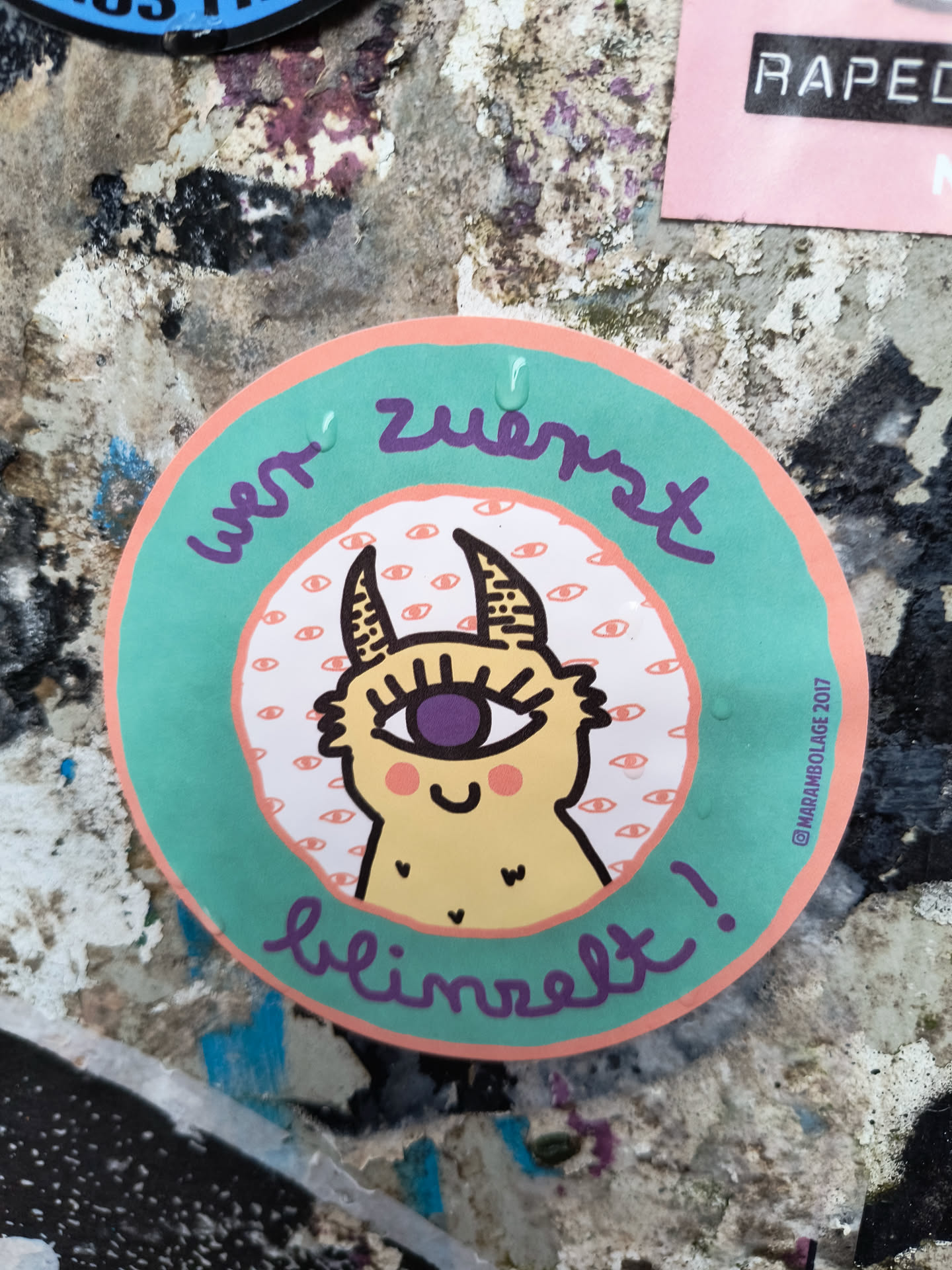 This round sticker showcases a cute, yellow monster with a single, large purple eye. The creature has small horns and a charming smile, set against a background pattern of numerous tiny eyes. The text 'Wer zuerst blinzelt!' curves around the design, written in a playful, handwritten style.