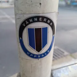 Street sticker A round sticker with a central shield-shaped logo. The shield is primarily blue with a vertical black stripe.  The words "VÄNNERNA" and "UPPSALA" encircle the shield in a circular black design.  The sticker appears to be affixed to a pole outdoors.