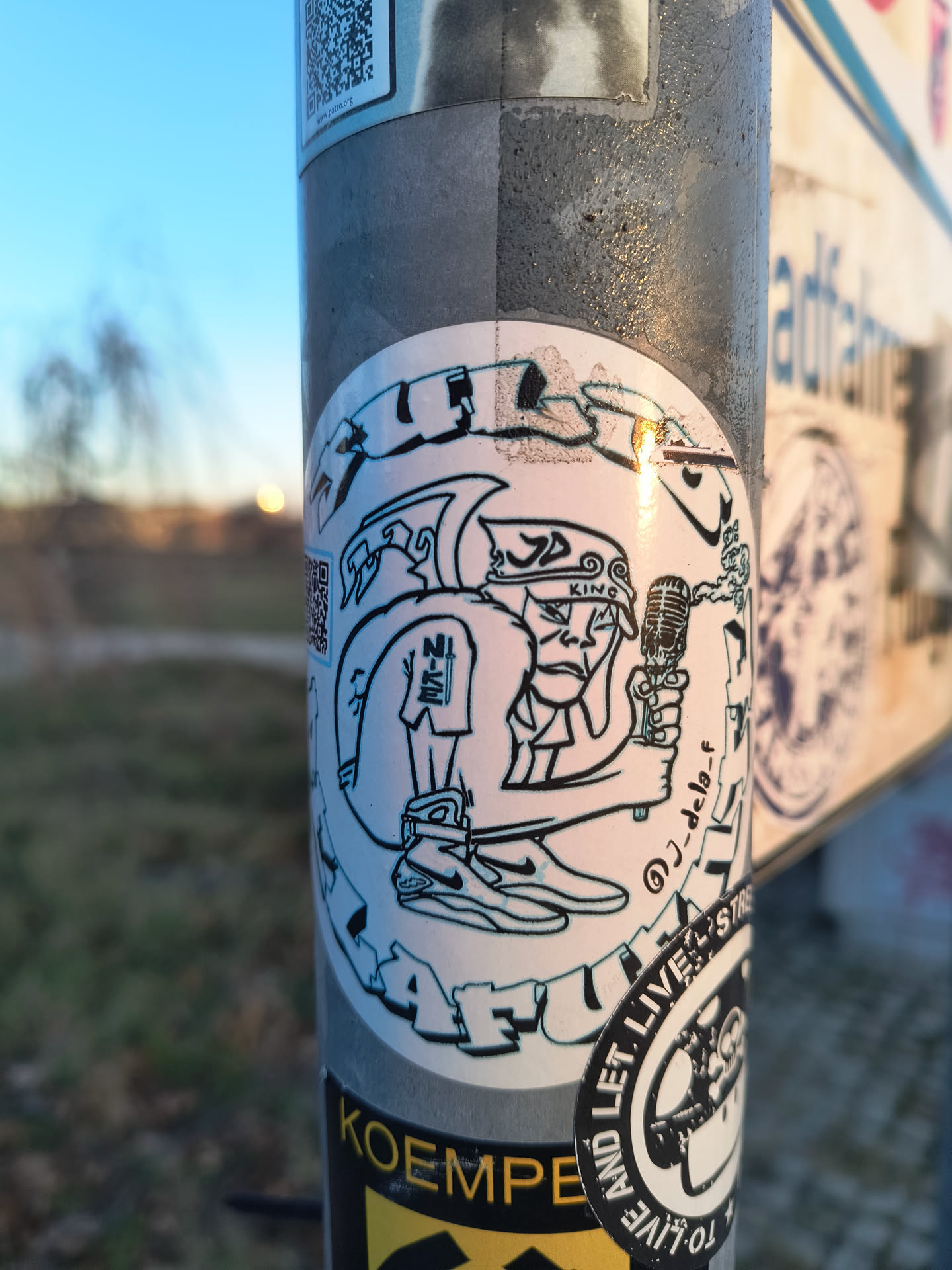Street sticker A circular sticker featuring a stylized, cartoonish male character in black line art on a white background. The character has a stern expression, a small beard, and wears a backward baseball cap with 'KING' written on it. He is holding an old-school microphone in his right hand, from which smoke or sound waves emanate. He wears shorts with 'NI-KE' on the left leg and prominent sneakers. The artist's handle '@J-dela-f' is visible on the right side of the character. The overall style is reminiscent of urban street art or graffiti.