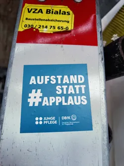 Street sticker A blue sticker promoting a movement with the slogan "Aufstand statt Applaus" (Rebellion instead of applause). It features the logos of Junge Pflege (Young Care) and DBfK (German Federation of Professionals in the Health and Welfare Sector).