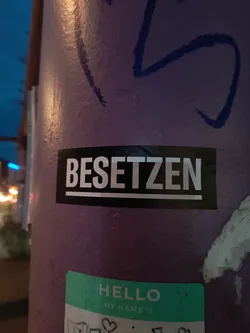Street sticker The sticker is rectangular with a black background and white, sans-serif text. The word 'BESETZEN' is centered on the sticker, underlined by a thin white line. The text is large and blocky, giving it a strong, impactful appearance. The sticker appears to be adhered to a purple, textured surface, possibly a pole, which also has some blue graffiti on it.