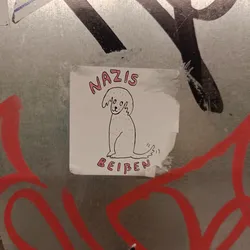 Street sticker A sticker with a drawing of a dog and the text "Nazis beißen" which translates to "Nazis bite". The sticker is placed on a wall with other graffiti.