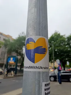 Street sticker A sticker expressing solidarity with Ukraine. It features a heart-shaped design with two hands embracing, representing support and unity. The colors of the Ukrainian flag are incorporated into the design.