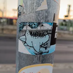 Street sticker A small sticker featuring a light blue bird illustration.  The bird is stylized and somewhat cartoonish. The background appears to depict a snowy or wintry scene. The sticker is affixed to a metal pole alongside other stickers.