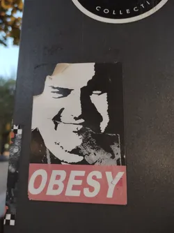 Street sticker A black and white stenciled portrait of a man with a smirk, overlaid with the word "OBESY" in pink letters. The sticker has a slightly damaged edge.