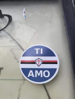 Street sticker A round sticker with a blue border and white center. It features the text 'TI AMO' separated by a band of red, white, black, and white with a shield containing a red cross. The color scheme and design are associated with the Italian football club Sampdoria.